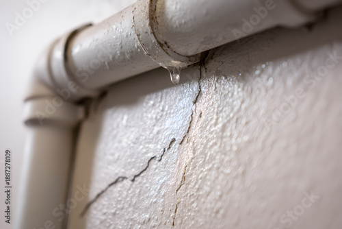 Water drips from a white pipe above a cracked wall in a residential building during daytime showing signs of damage