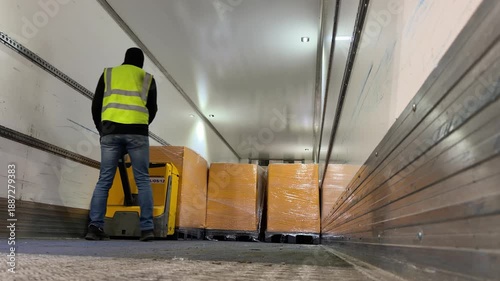 Warehouse operative unloading orange-wrapped pallets from a truck trailer using an electric ride-on pallet jack