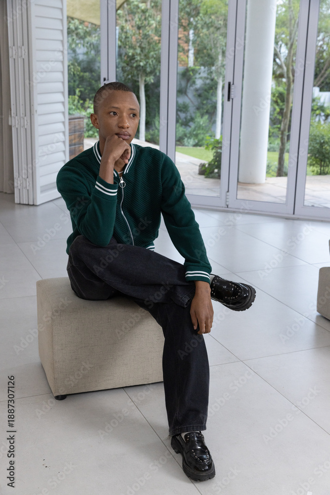 Naklejka premium African American man sitting on beige ottoman in modern living room wearing dark green sweater