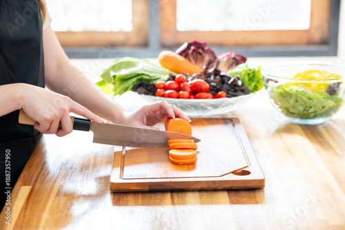 An Asian woman is chopping carrots on a kitchen table to make a salad. The concept of healthy eating and dieting is conveyed.