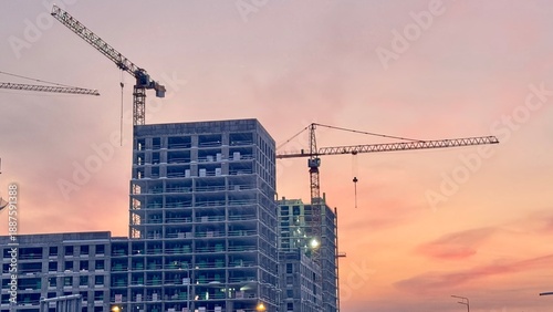 Tableau sur toile Construction site with towering cranes and multi-story residential buildings under development against a vibrant sunset or sunrise sky