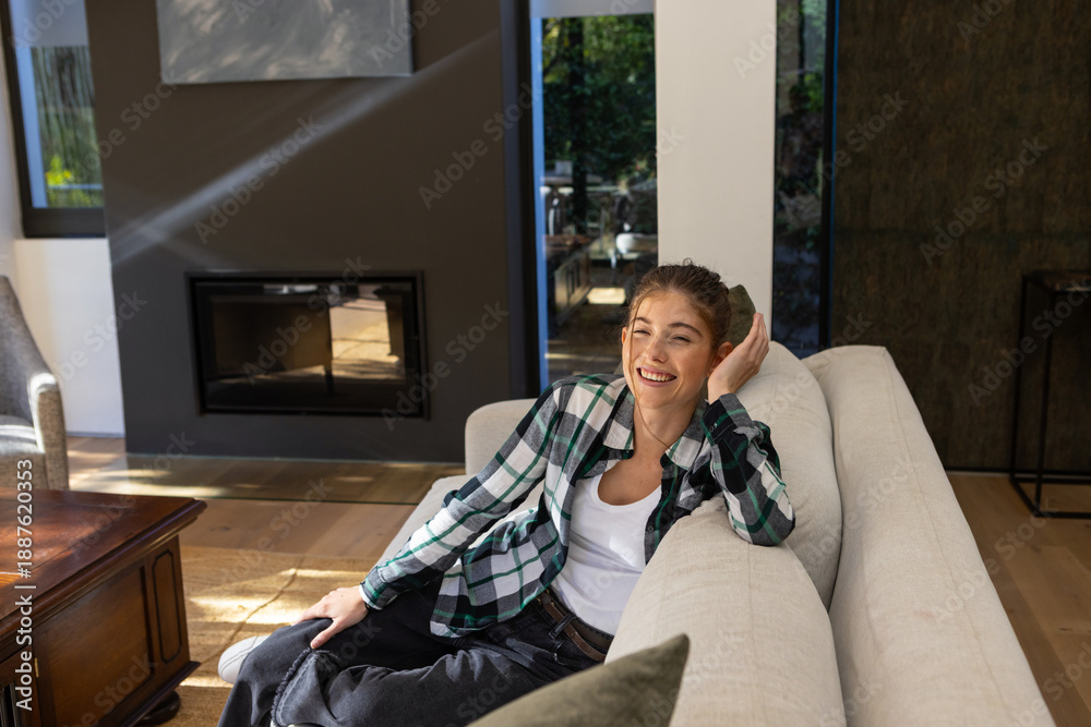 Fototapeta premium Woman reclining on beige sofa and smiling in living room with fireplace, copy space