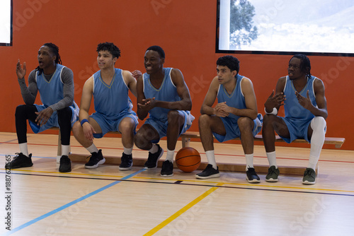 Diverse male teammates in blue uniforms resting on wooden bench on sports hall gym court with ball