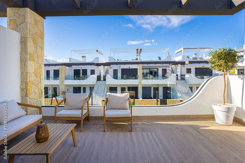 Fototapeta premium Sunny modern apartment balcony with wood furniture and view of white minimalist buildings