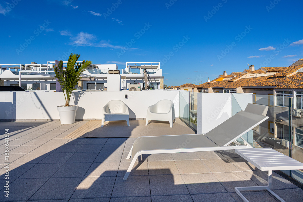 Fototapeta premium Modern rooftop lounge with white sunbeds and distant tiled rooftops under blue sky