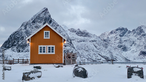 Fotografie A traditional yellow rorbu cabin at Sakrisøy village in Lofoten, Norway, with snow capped mountains in winter