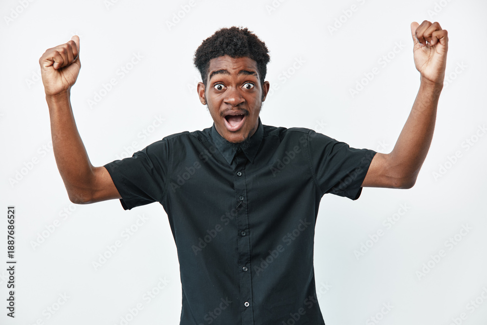 Fototapeta premium Young black man with dark skin wearing black shirt raising arms and expressing excitement or surprise on white background. Portrait of emotional african male.