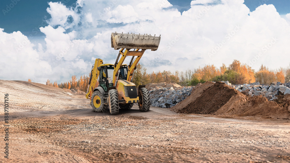 Fototapeta premium A loader lifts material at a construction site surrounded by piles of earth and rocks with trees in the background during daylight hours