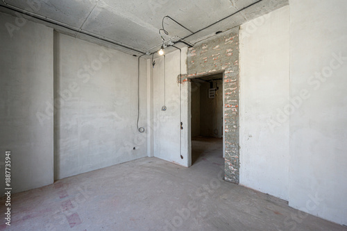 Unfinished room with plastered walls, exposed ceiling pipes, rough concrete floor, visible electrical wiring, open doorway, minimal fixtures, awaiting final renovation.