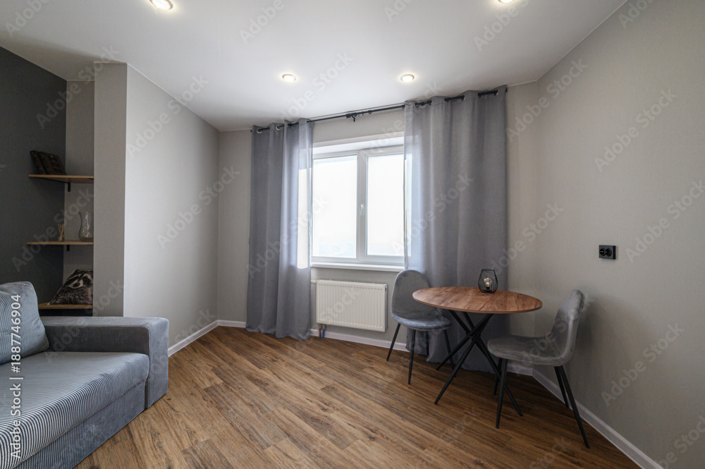 Fototapeta premium cozy modern room with a grey sofa, wooden flooring, a round dining table with two chairs, grey curtains, built-in shelves, recessed ceiling lights, and a large window with natural light.