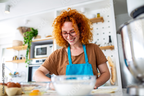 Billede på lærred Happy redhead woman in blue apron kneading dough on a kitchen counter