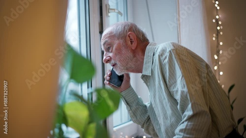 Old man standing by window, making phone call.