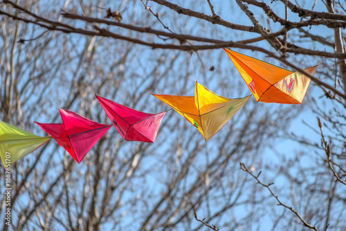 Colorful kites in red, yellow, and orange flying against clear blue sky with bare tree branches, capturing joyful outdoor kite festival atmosphere
