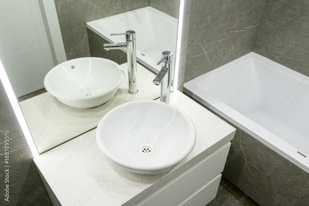 Fototapeta premium Modern bathroom with two round vessel sinks, chrome faucets, white countertop, LED-lit mirror, grey marble tiles, sleek bathtub, minimalist design.