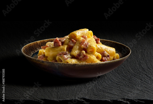 Dark bowl with rigatoni pasta carbonara on black stone
