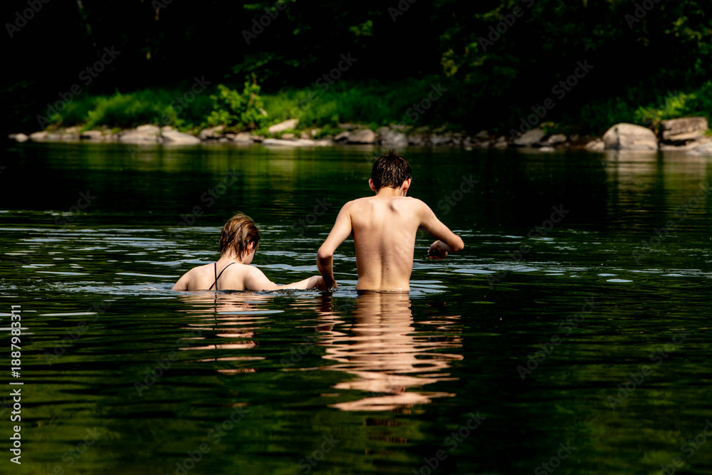 Fototapeta premium Back view of two people wading and swimming in deep natural river or lake with dark green water and rocky forested shoreline. 