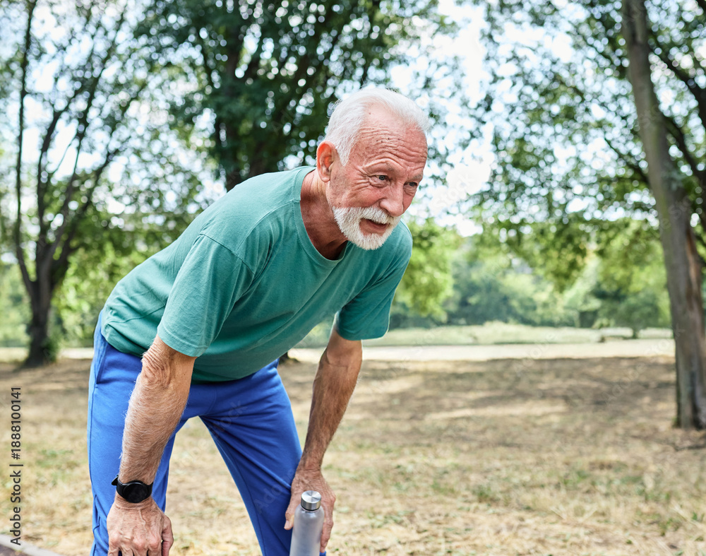 Fototapeta premium Portrait of a mature senior man exercising in a park, tired mature male running and taking a break holding a water bottle, outdoors, healthy lifestyle and cardio exercise in nature concepts, vitality 