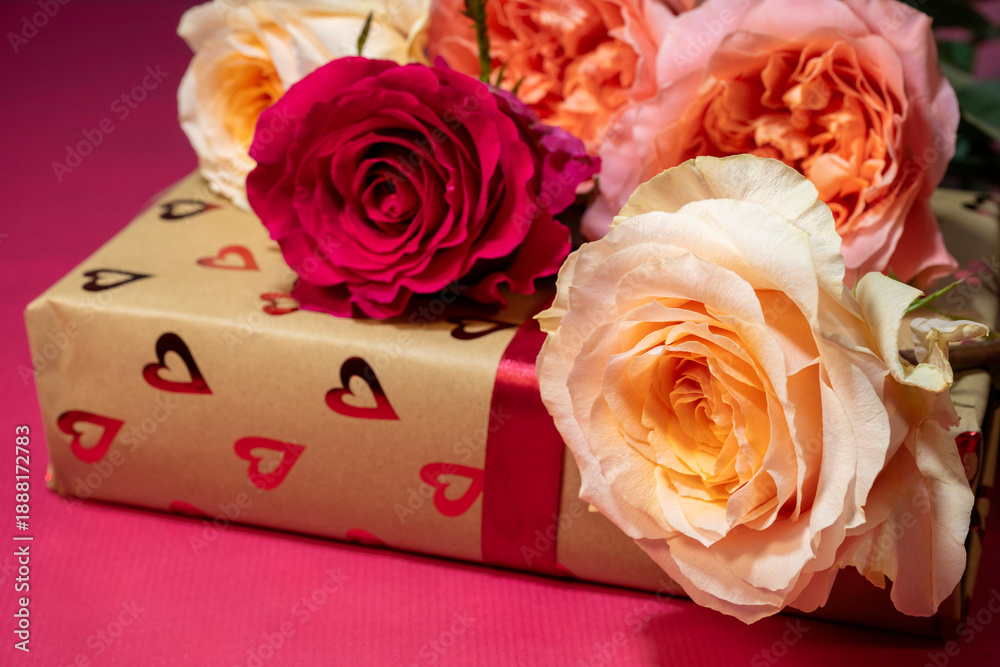 Fototapeta premium a gold gift box wrapped in heart-patterned paper and a red ribbon, topped with a blooming red rose and several cream and peach roses on a pink background.