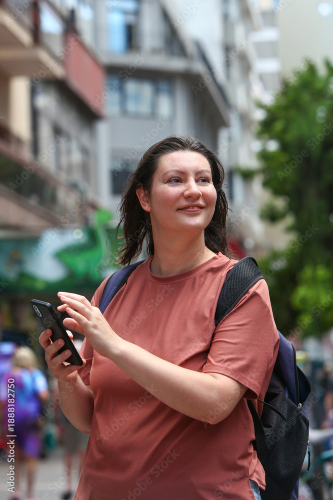 Φωτογραφία urban lifestyle image of an adult woman walking on a busy city street, smiling while holding a smartphone and wearing a backpack