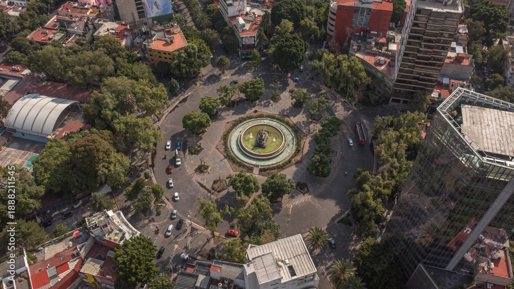 Fototapeta premium Aerial view of the Fuente de Cibeles roundabout in the Roma neighborhood of Mexico City. Capturing the iconic fountain surrounded by tree-lined streets, urban traffic, and residential buildings.