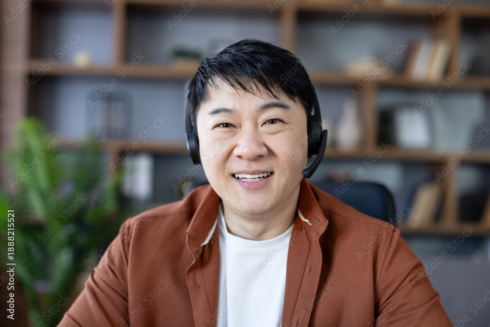 Fototapeta premium Asian man smiling, wearing a headset with a microphone, participating in an online video conference or remote consultation from a home office environment
