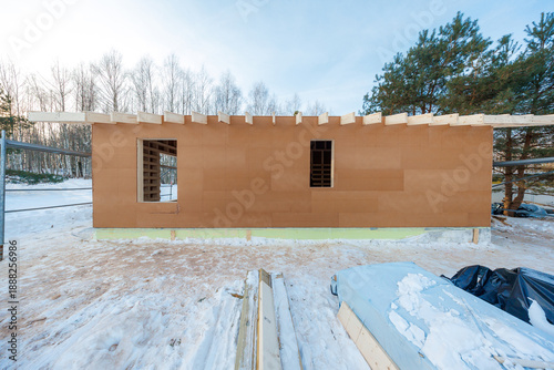 Single story wooden structure under construction in winter light, with exposed roof joists, plywood sheathing, window openings, studs, birch trees, pines, and insulation.