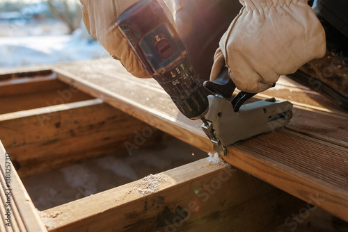 Worker in insulated gloves uses a cordless drill and flooring clamp to fasten grooved timber boards onto joists, frost visible, snow present, long warm sunlight shadows.