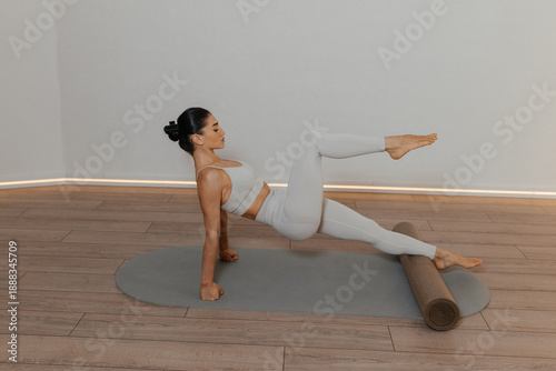Pilates foam roller. Athletic brunette woman practices pilates on foam roller wearing light grey outfit on grey mat. Controlled exercises show strength flexibility balance and body control.