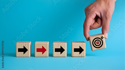Hand placing a wooden block with a target on it at the end of a line of blocks with arrows, representing achieving a goal