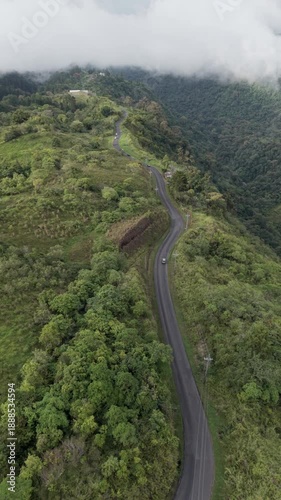 Drone following car along winding mountain road on steep ridge with dramatic green mountains and low clouds