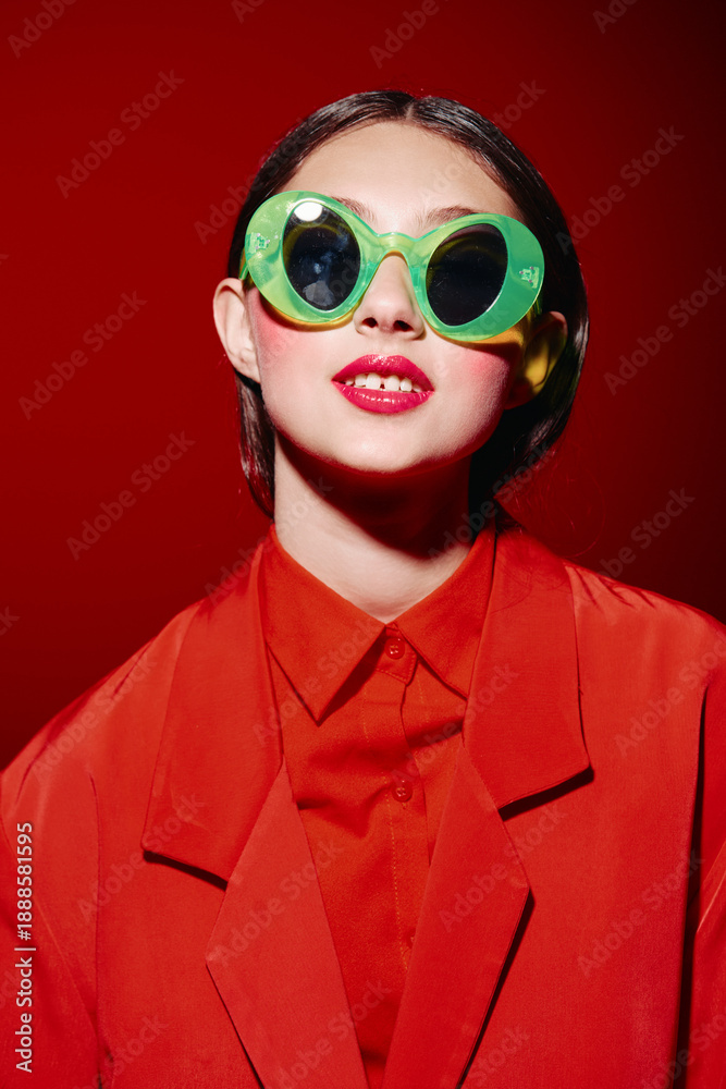Fototapeta premium Stylish woman with bright red lipstick wearing oversized green sunglasses and a red outfit against a vibrant red background. Concept of beauty, fashion, creative style, and modern trends.
