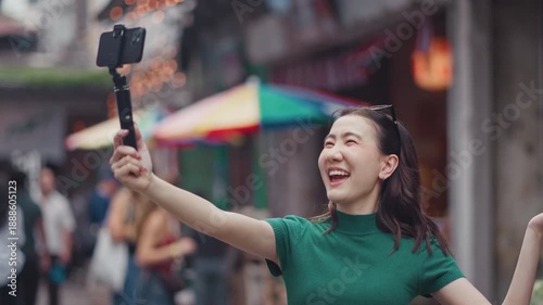 Happy young Asian woman vlogger recording a travel video with a smartphone on a selfie stick while exploring a city street. Modern influencer and digital lifestyle concept.