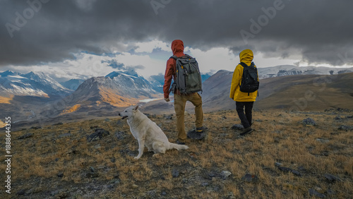 Quadro su tela A couple in bright rain jackets and a white dog stand on a mountain ridge during an autumn hiking trip, looking at a dramatic landscape of distant snow-capped peaks under heavy, cloudy skies