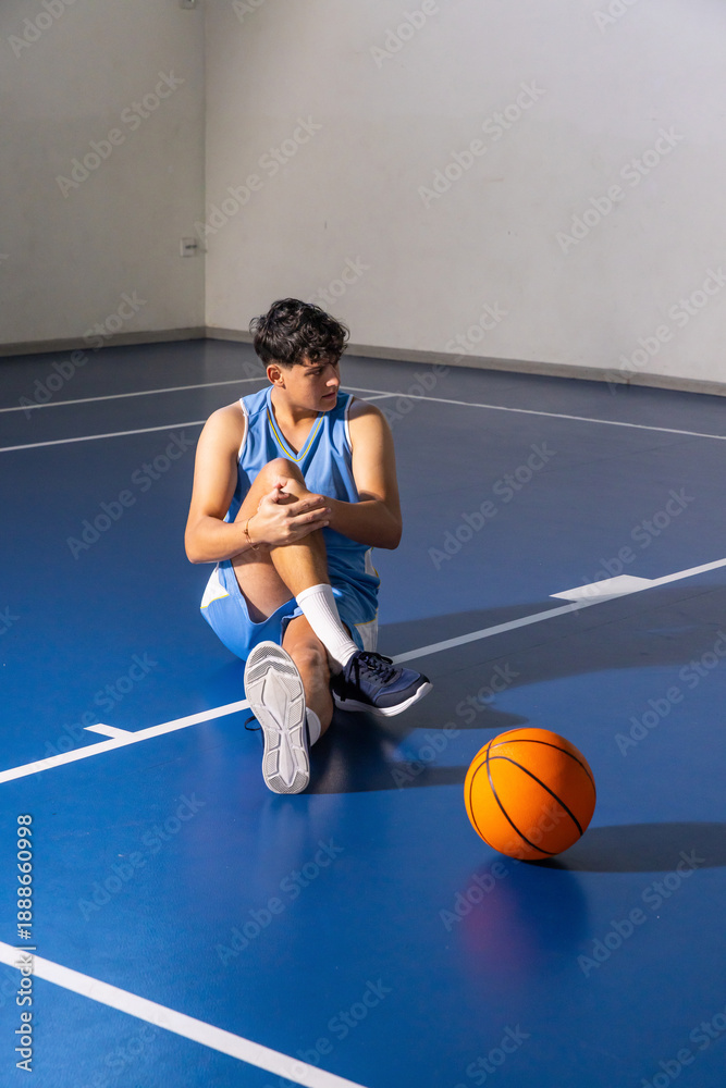 Naklejka premium Asian teenage male basketball player holding knee and stretching leg on court near orange ball