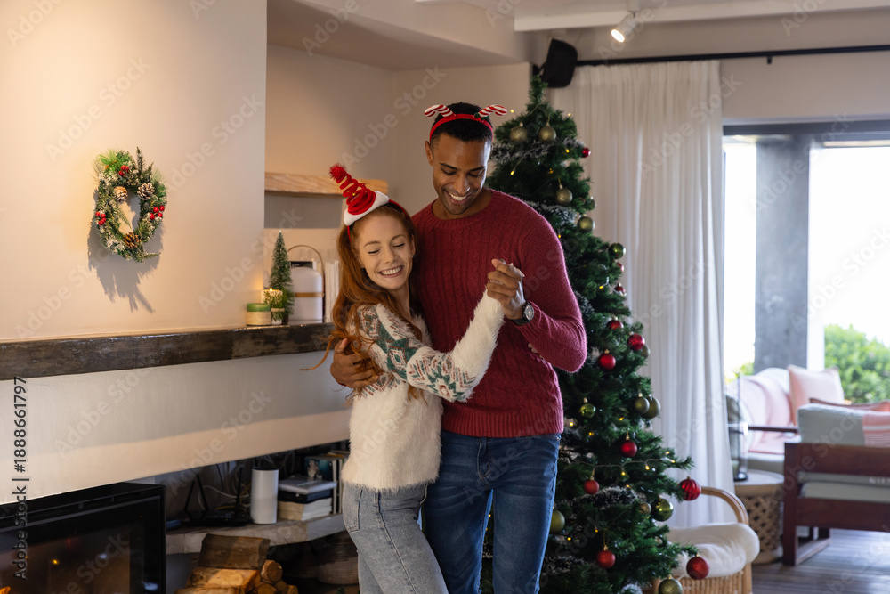 Naklejka premium Diverse couple dancing in fuzzy holiday sweaters in living room next to christmas tree, fireplace