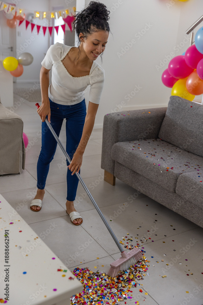 Fototapeta premium Teenage girl sweeping confetti with broom by gray couch in living room with balloons