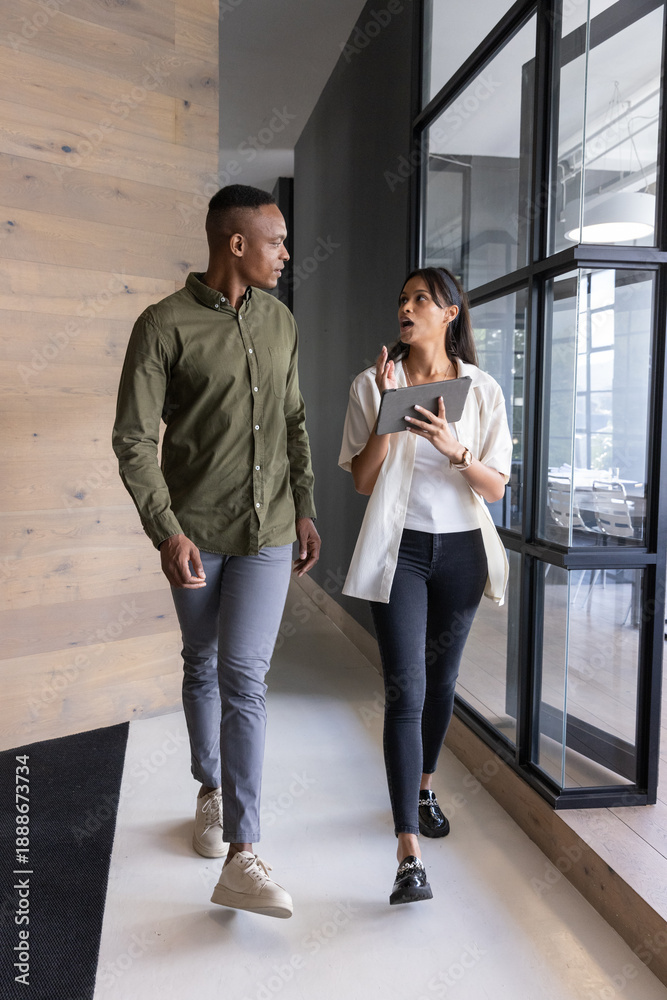 Fototapeta premium Diverse colleagues walking together conversing in office hallway holding tablet