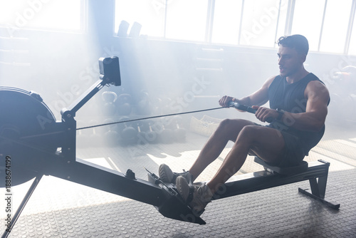 Fotografi Muscular adult man working on rowing machine near kettlebells in gym under sunli