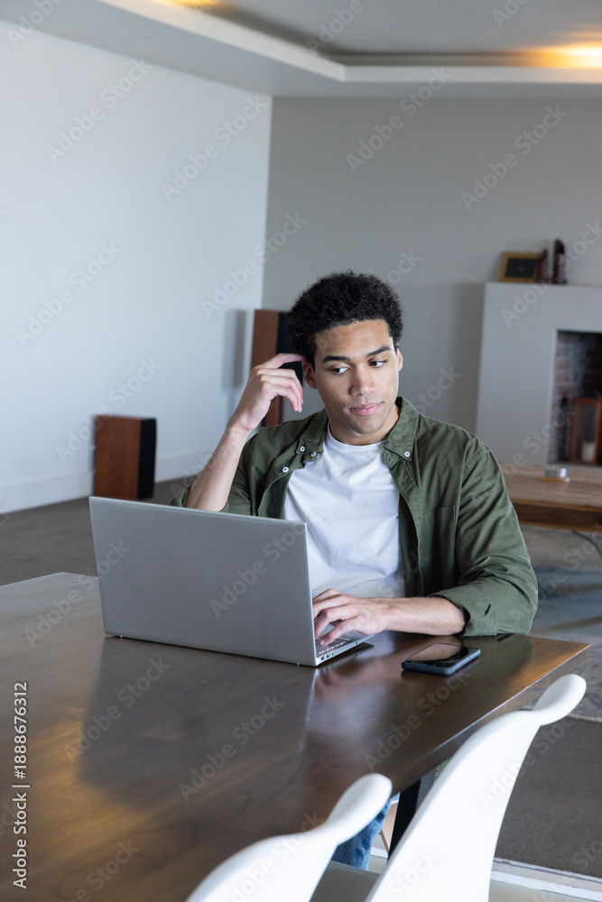 Fototapeta premium African american man sitting at dining table using laptop with smartphone in living room