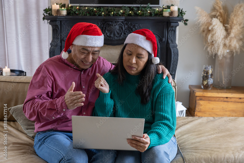 Obraz premium Asian couple with laptop and pointing at screen wearing santa hats in living room