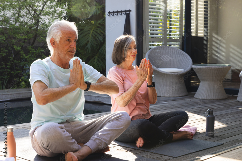 Fototapeta premium Senior couple meditating while holding hands on yoga mats on wooden poolside deck