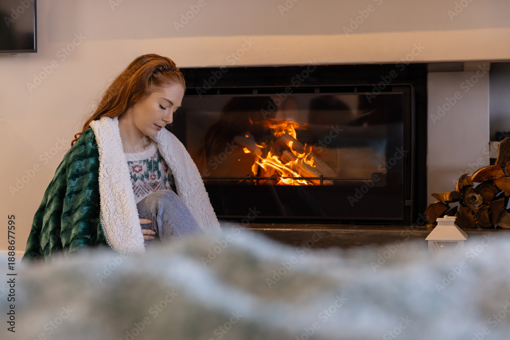 Obraz premium Red-haired woman sitting on floor by glass fireplace in living room in plush blanket, copy space