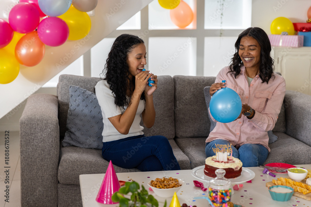Fototapeta premium Diverse mom and teenage daughter inflating blue balloons in living room with birthday cake