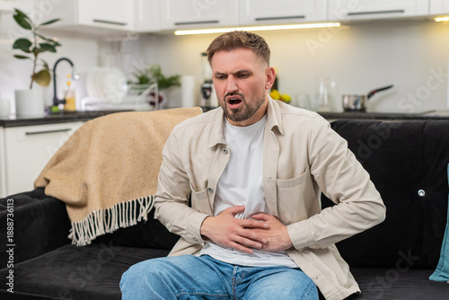 Young man at home presses hands to stomach feels cramps and pain after heavy fatty food and breathes. Caucasian guy on sofa curls slightly and shows nausea spasm and low mood while waiting for relief.