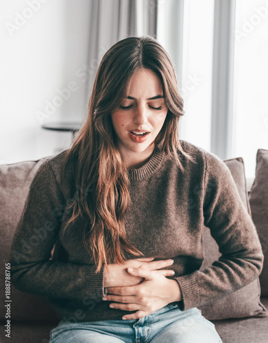 Foto Young woman with stomach pain sitting on the couch alone at home, holding her stomach, touching and massaging her abdomen