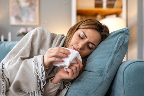 Sick young woman lying on sofa at home blowing nose with paper tissue while suffering from severe cold flu or allergic rhinitis symptoms under a warm blanket healthcare and medical