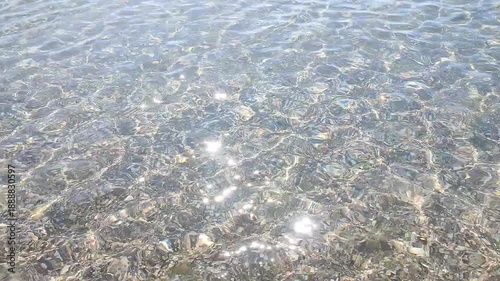 Clean transparent water on shallow sandy seashore with shells with shells on sunny summer day. Sun glare on surface of water. Small waves on sea coast. Nature backdrop. Natural background.