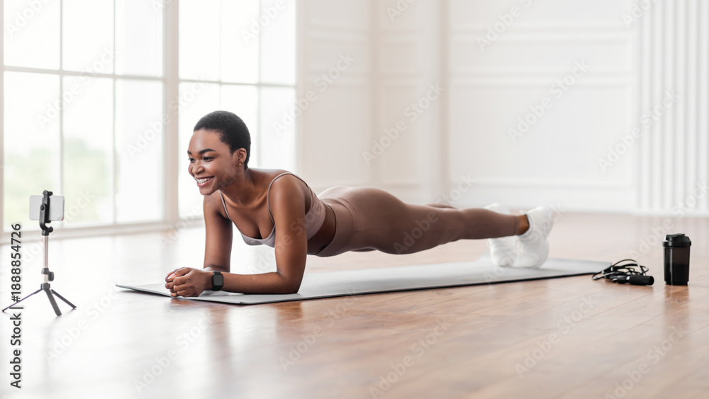 Fototapeta premium Joyful black lady in sportswear planking in living room, watching fitness video or having online conference with fitness trainer, sporty woman exercising and looking at smartphone screen, free space