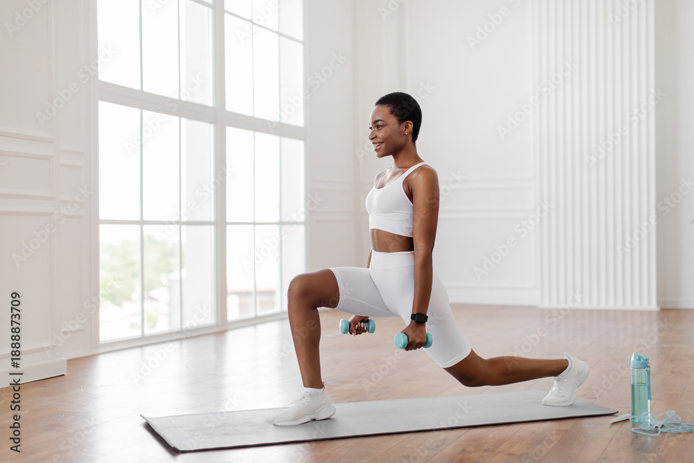 Fototapeta premium Sport And Healthy Lifestyle. Side profile view of smiling fit African American woman doing forward walking lunges with dumbbells, exercising at home. Smiling lady training on yoga mat near window