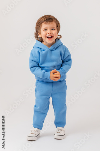 Vertical full-length portrait of a 3-year-old boy who is laughing cheerfully and looking at the camera. He is wearing a blue hoodie, pants, and sneakers.Photo